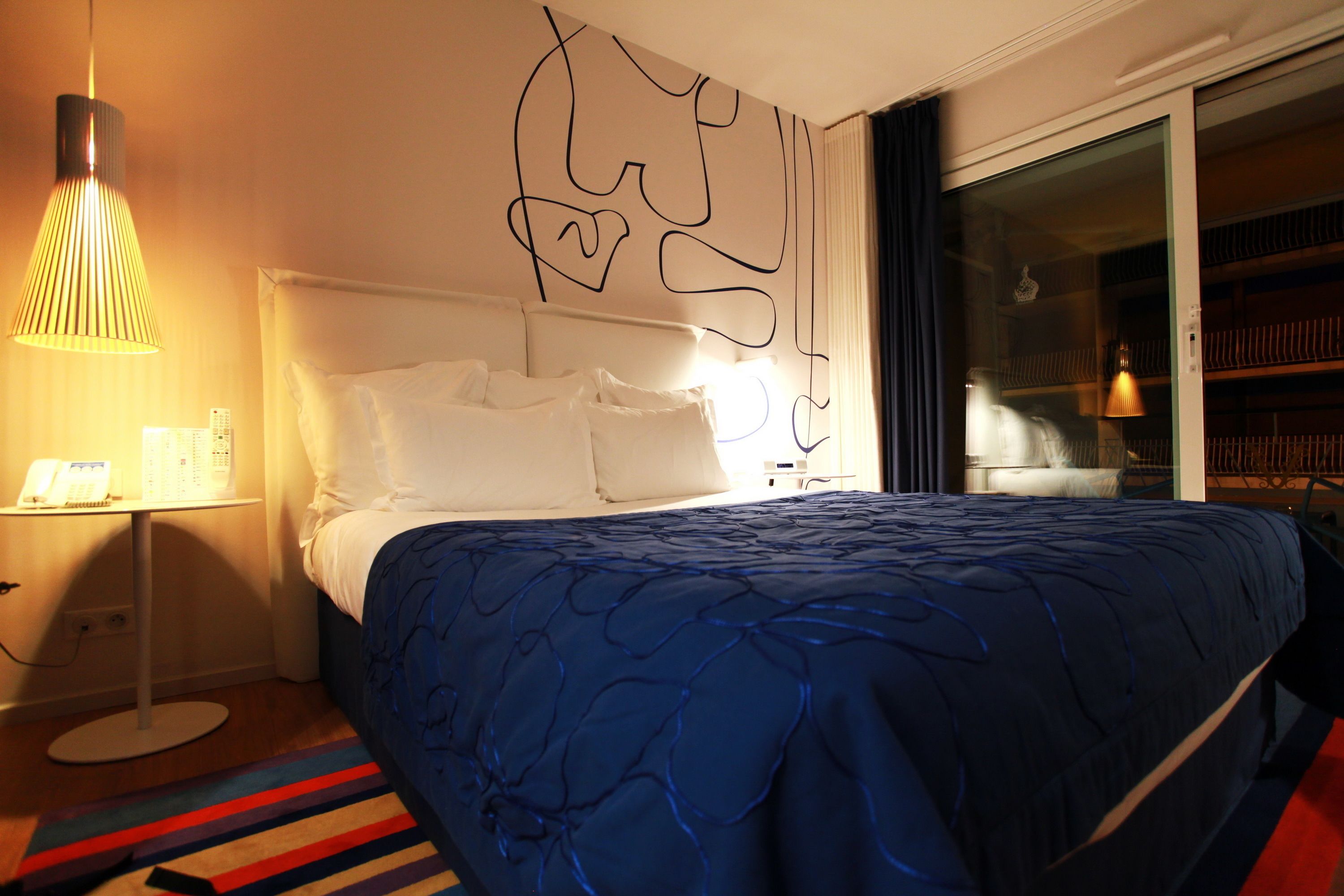 Habitación de hotel con una cama, una mesita de noche con lámpara y vistas a un balcón con estanterías.