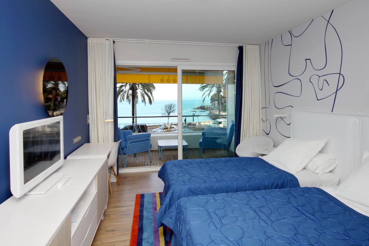 Une chambre d’hôtel avec deux lits, une télévision, un balcon avec vue sur la mer et un coin repas comprenant des chaises et une table.