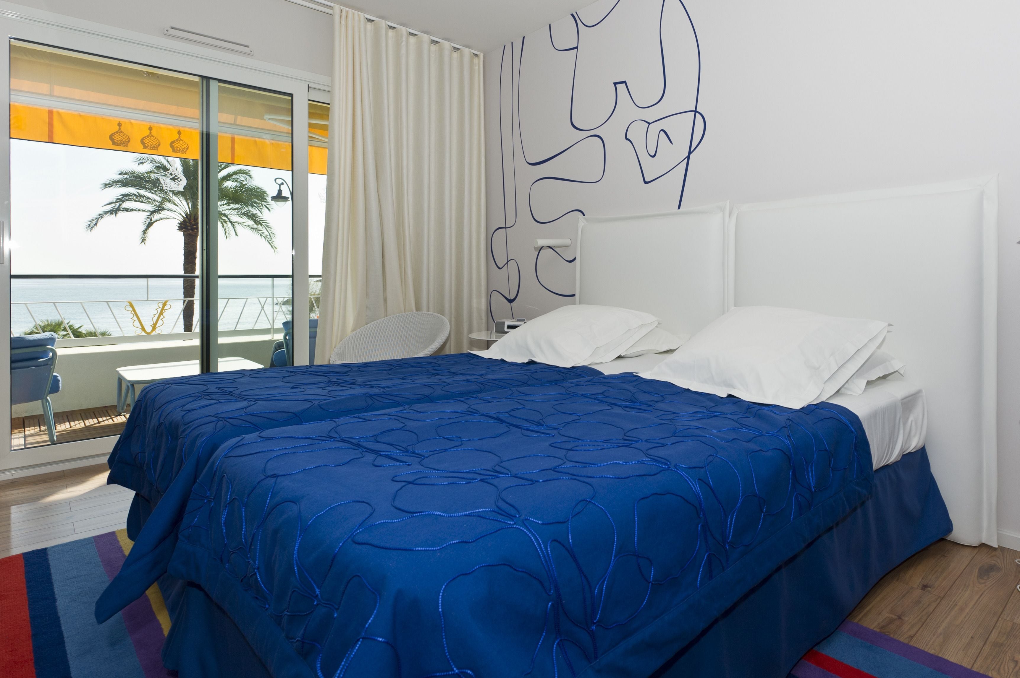 A hotel room with a large bed, a view of the sea, a balcony with a chair, and a wall with abstract art.