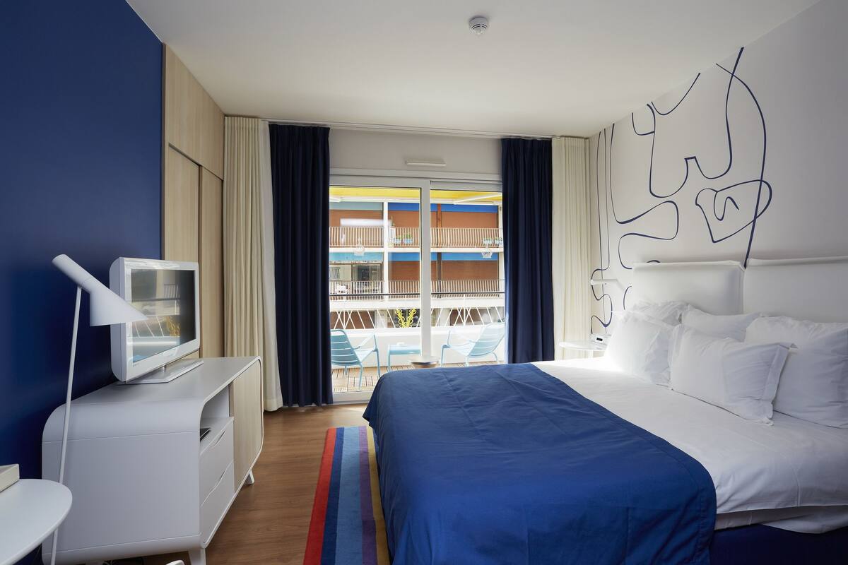 Une chambre d’hôtel comprenant un lit, un téléviseur posé sur un meuble, un bureau et une vue sur un balcon avec des sièges d’extérieur.
