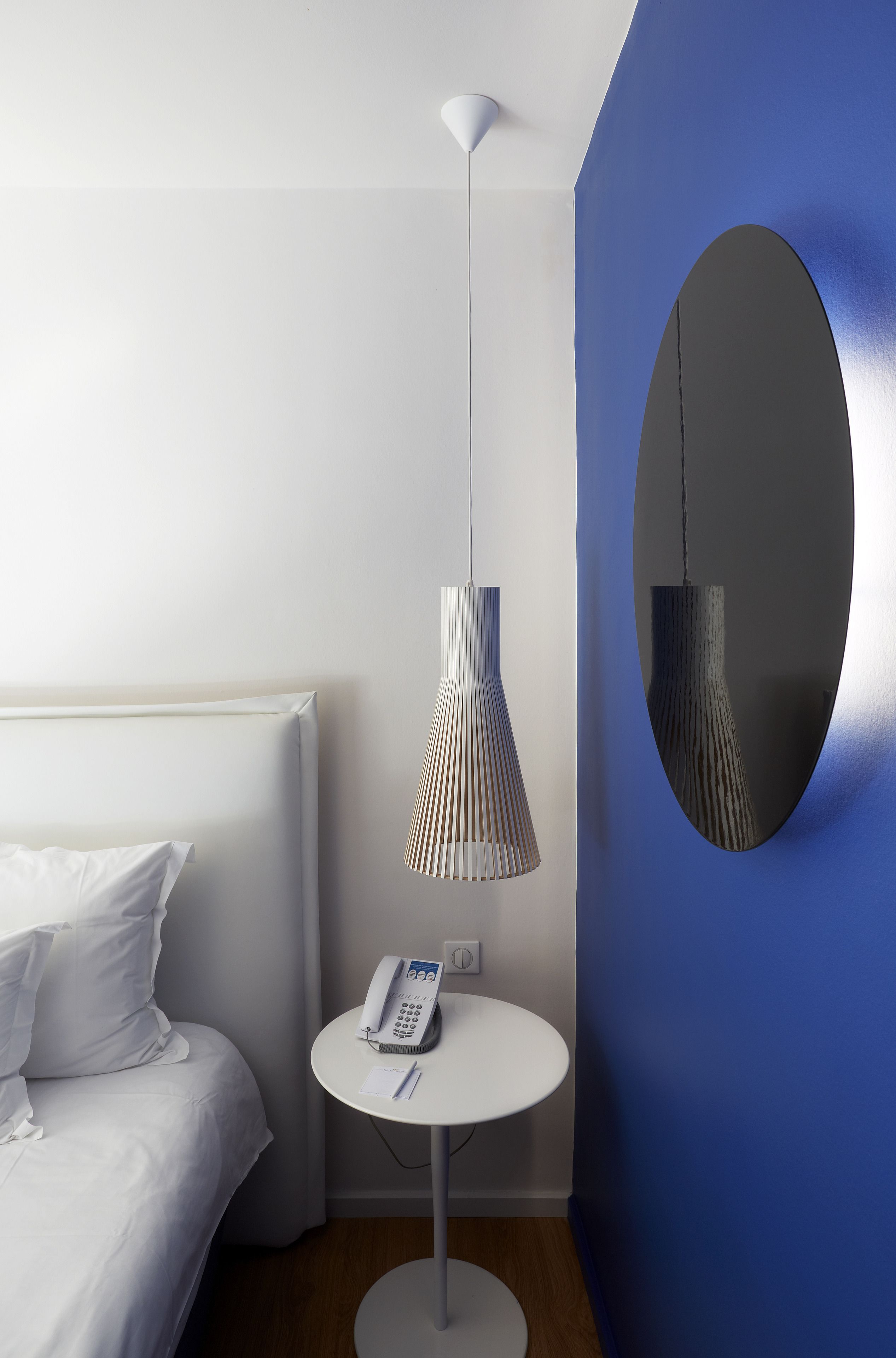 Una habitación de hotel con cama, mesita de noche con teléfono, lámpara y espejo en la pared.