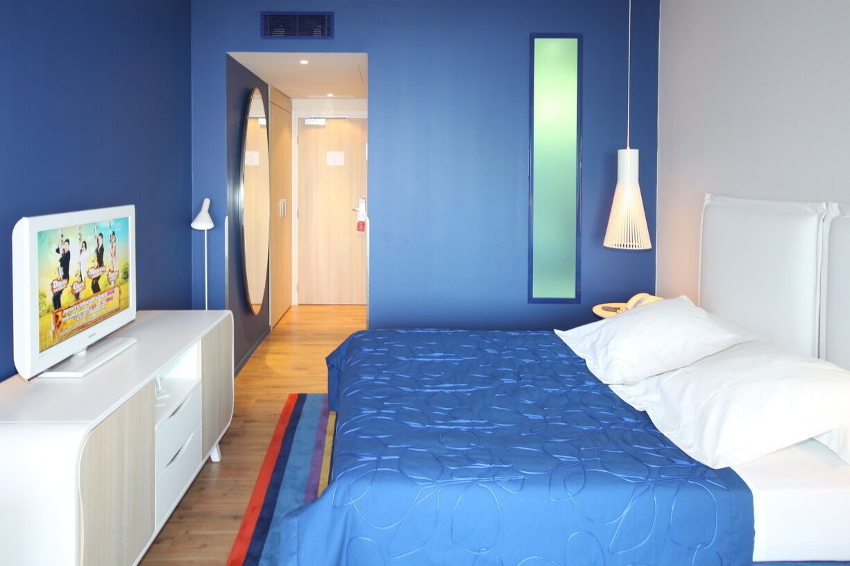 Een slaapkamer met een bed, een televisie op een witte kast, een spiegel en een deur die naar een andere kamer leidt.