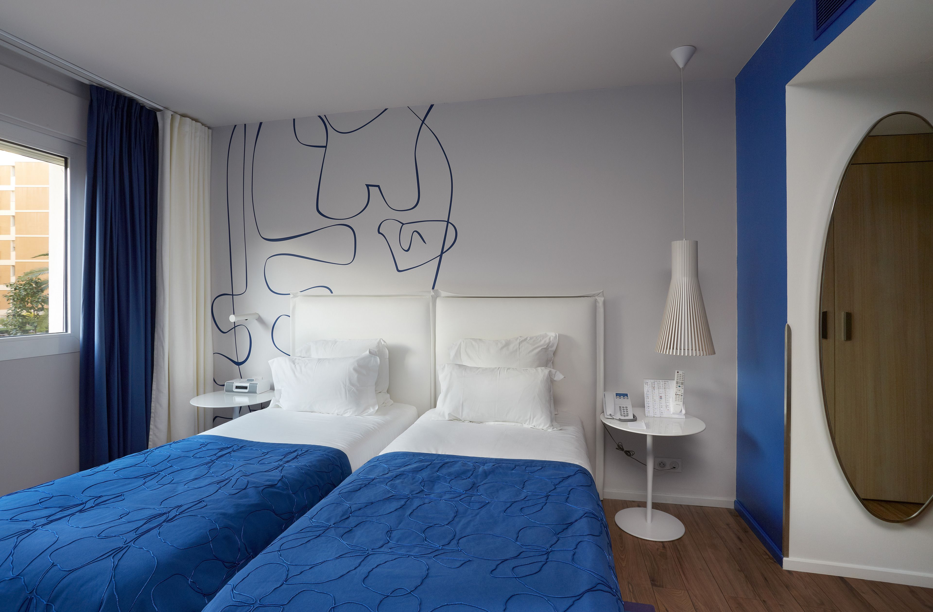 A hotel room with two beds, a small table with a lamp, and a large window with curtains.