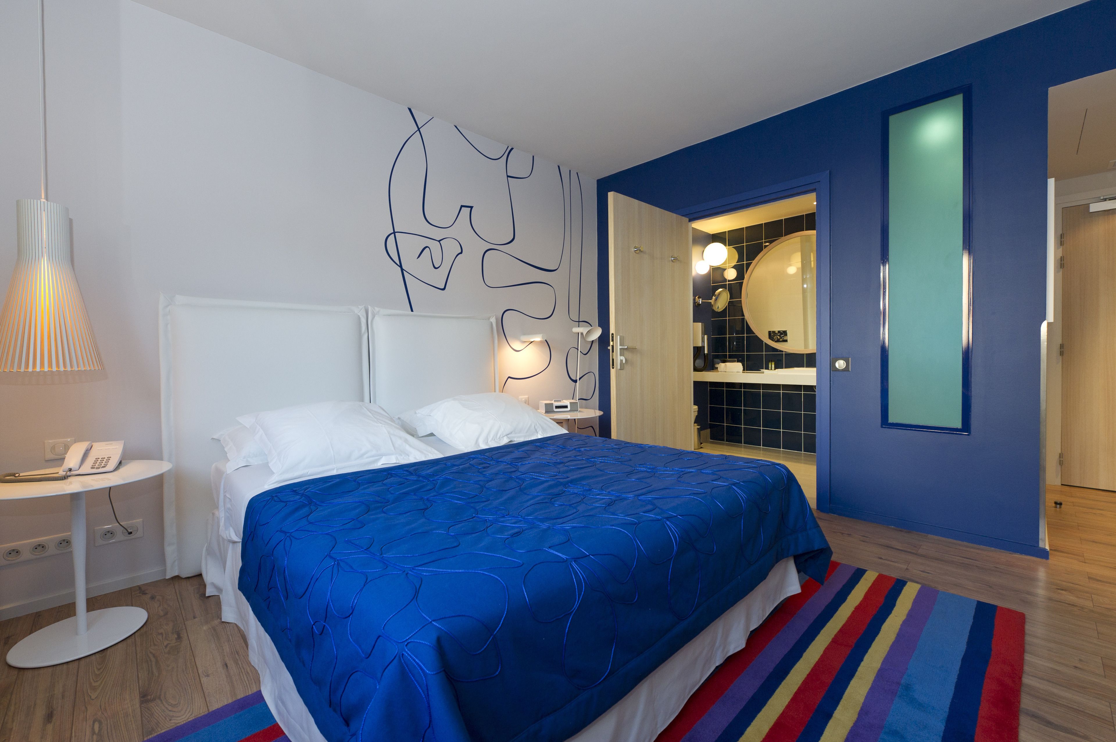 Un dormitorio moderno con una cama grande, una mesita de noche con un teléfono, un espejo y un baño visible a través de una puerta abierta.