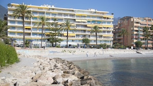 Un hôtel à plusieurs étages, doté de balcons, situé près d’une côte rocheuse bordée de palmiers.