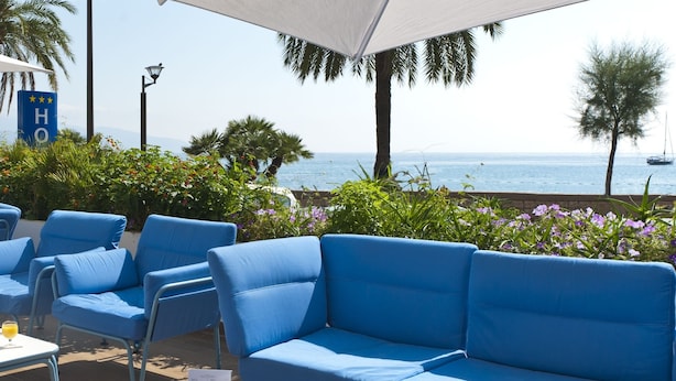 Une terrasse en bord de mer avec des sièges bleus, une table avec des boissons et une vue sur la mer et les montagnes.