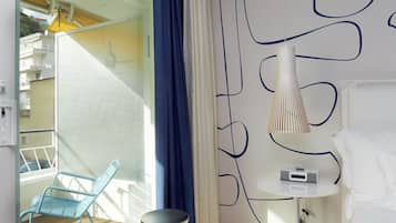 A modern hotel room with a glass door leading to a balcony, a blue chair, a small round table, a bedside lamp, and a colorful striped rug.