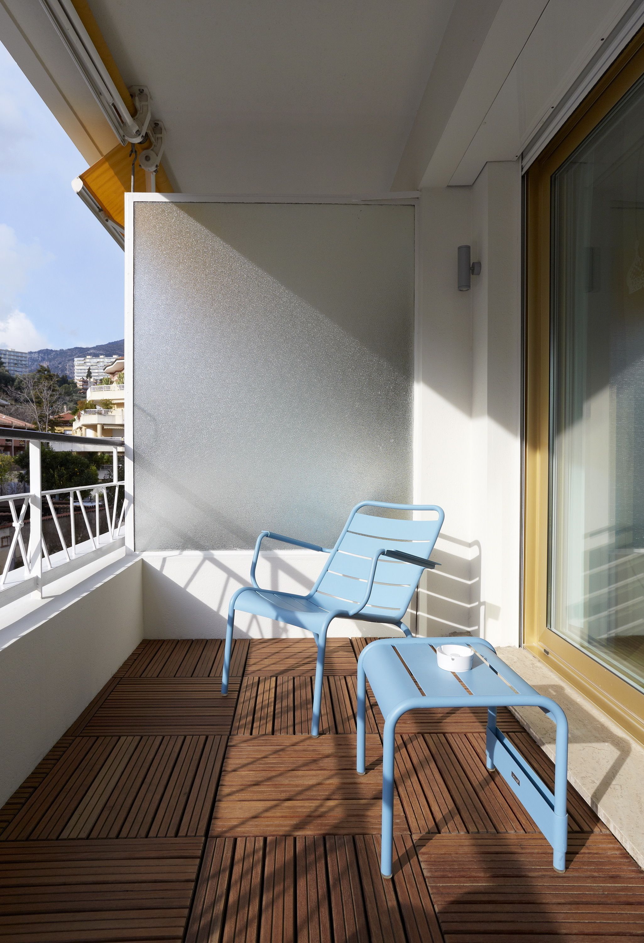 A balcony with two blue chairs and a small table, wooden flooring, and a view of buildings and hills.