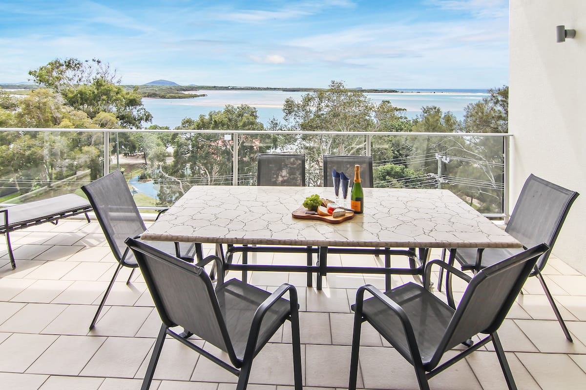 A balcony with a table set for two, overlooking a scenic view of a body of water and distant land.