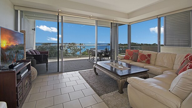 A modern living room with a large sliding glass door leading to a balcony, a flat-screen TV, a leather sofa, and a dining area with a table and chairs. - Space Holiday Apartments (Maroochydore)