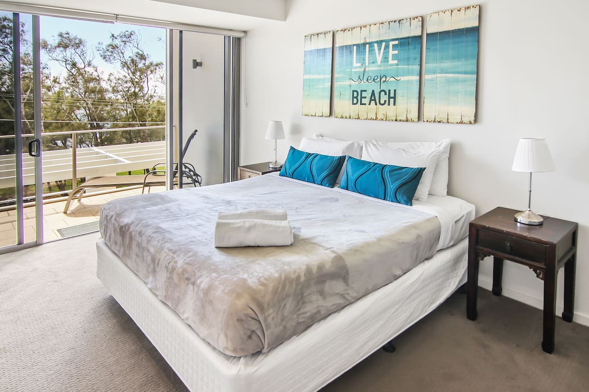 A hotel room with a large bed, a view of the outdoors, and a 'Live Sleep Beach' sign on the wall.