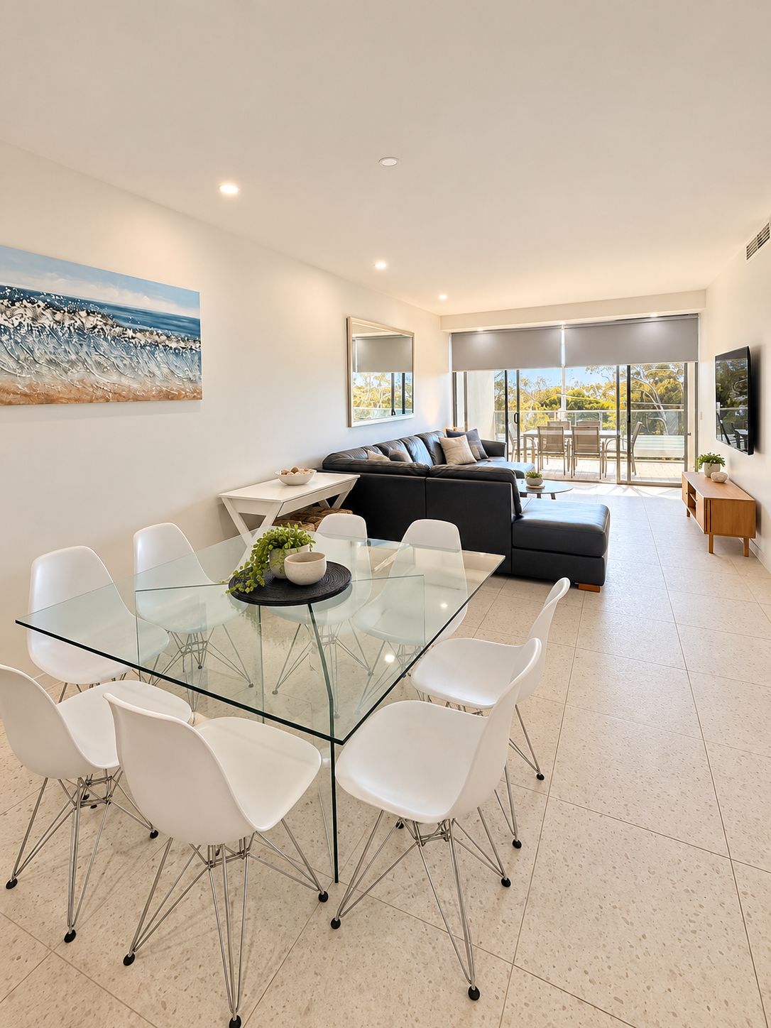 A modern living room with a glass dining table, white chairs, a sofa, and a large painting on the wall.