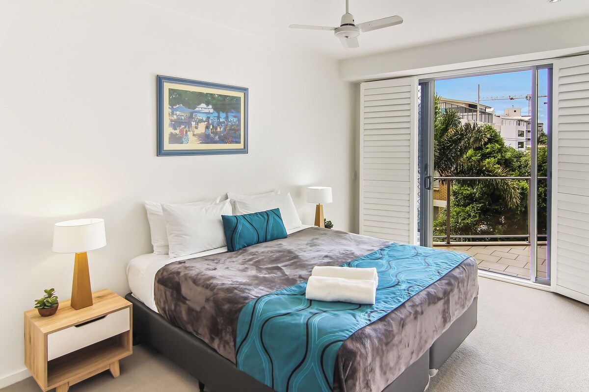 A bedroom with a bed, bedside tables, a ceiling fan, a window with a view, and a framed picture on the wall.
