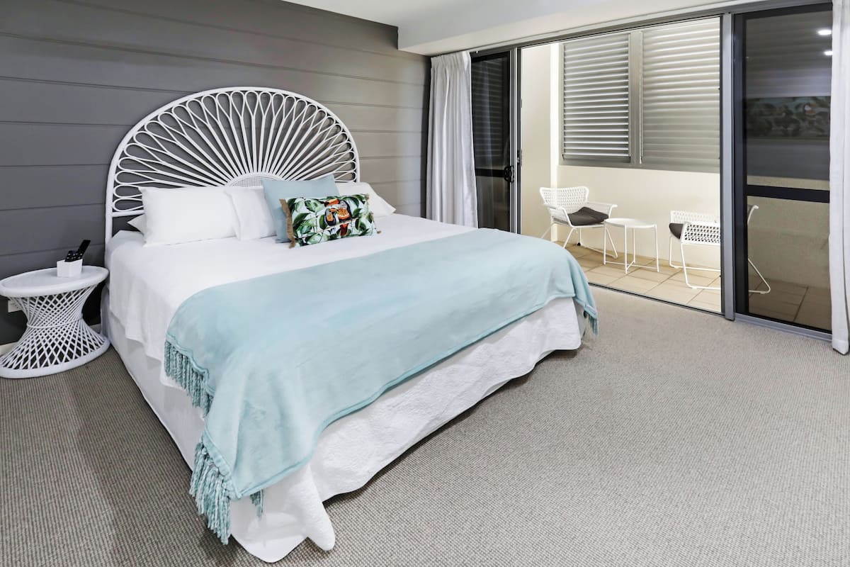 A bedroom with a bed, a side table, a white chair, and a balcony with a glass door.