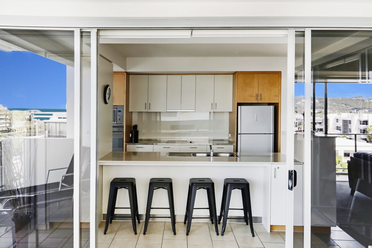 A modern kitchen with white cabinets, a stainless steel refrigerator, and a bar area with stools.