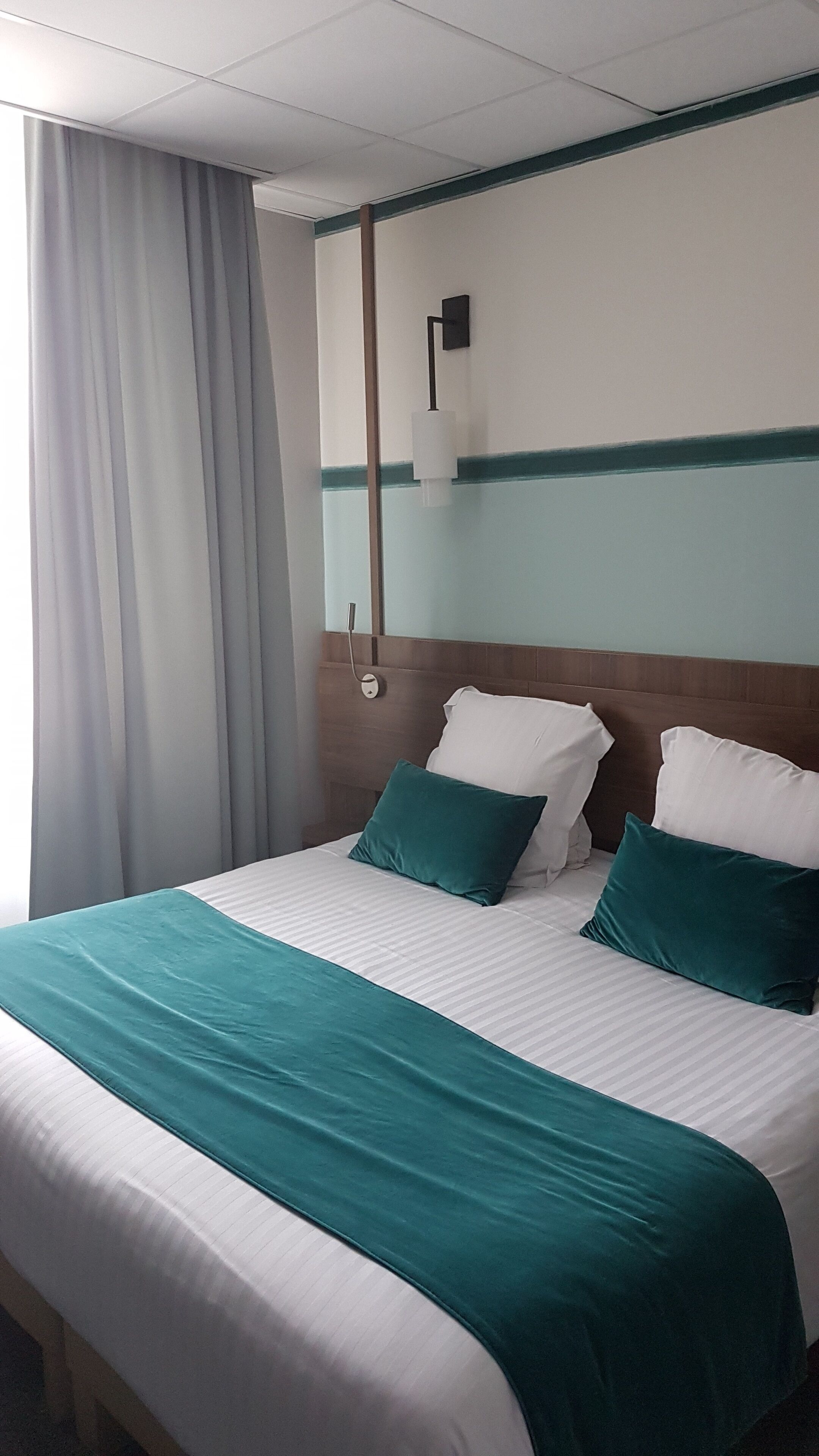 A hotel room with a bed, curtains, a wall-mounted light, and a wooden headboard.