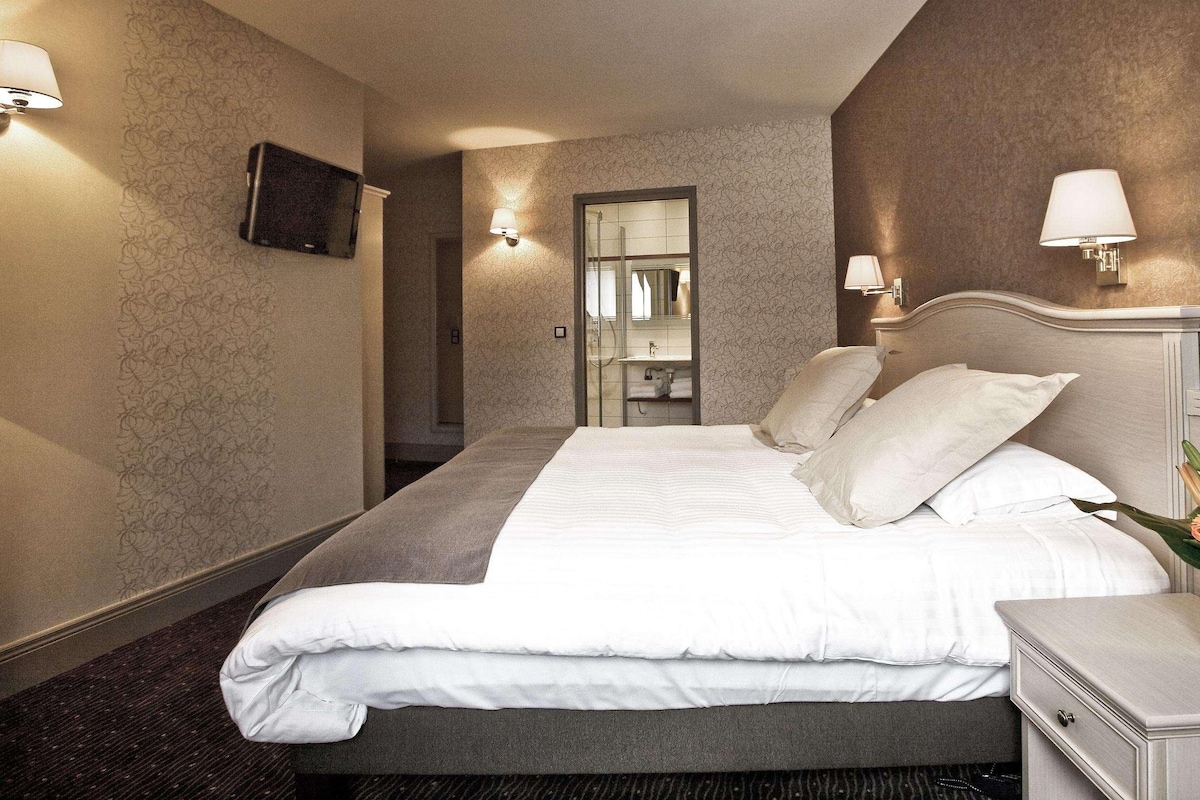 A hotel room with a large bed, two bedside lamps, a nightstand with a vase of flowers, and a television mounted on the wall.