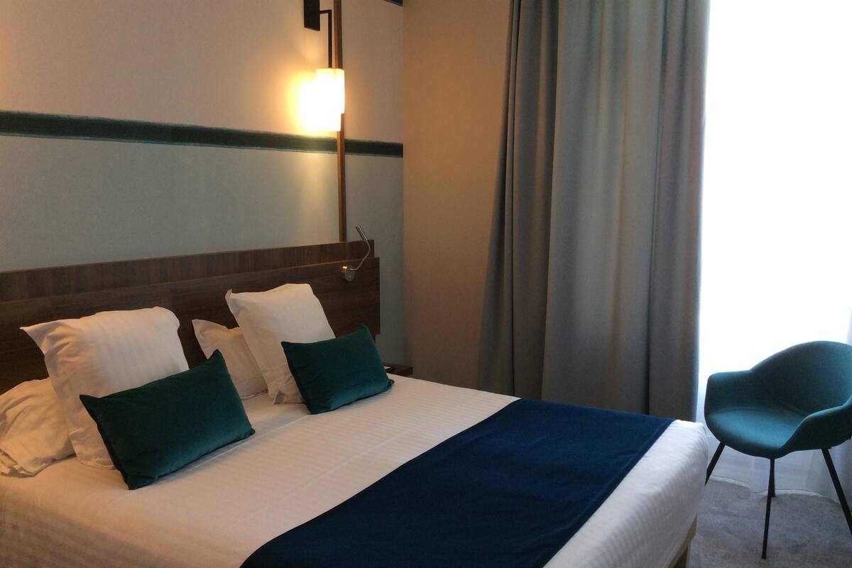 A hotel room with a bed, pillows, a nightstand, a lamp, a chair, and a window with curtains.