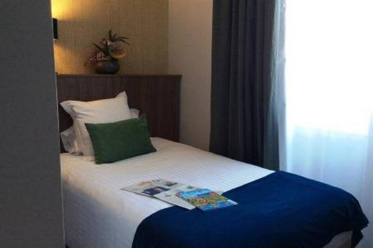 A hotel room with a bed, a nightstand with a lamp, a window with curtains, and a small potted plant.