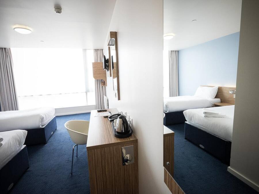A hotel room with two beds, a desk, a chair, a minibar, and a large window.