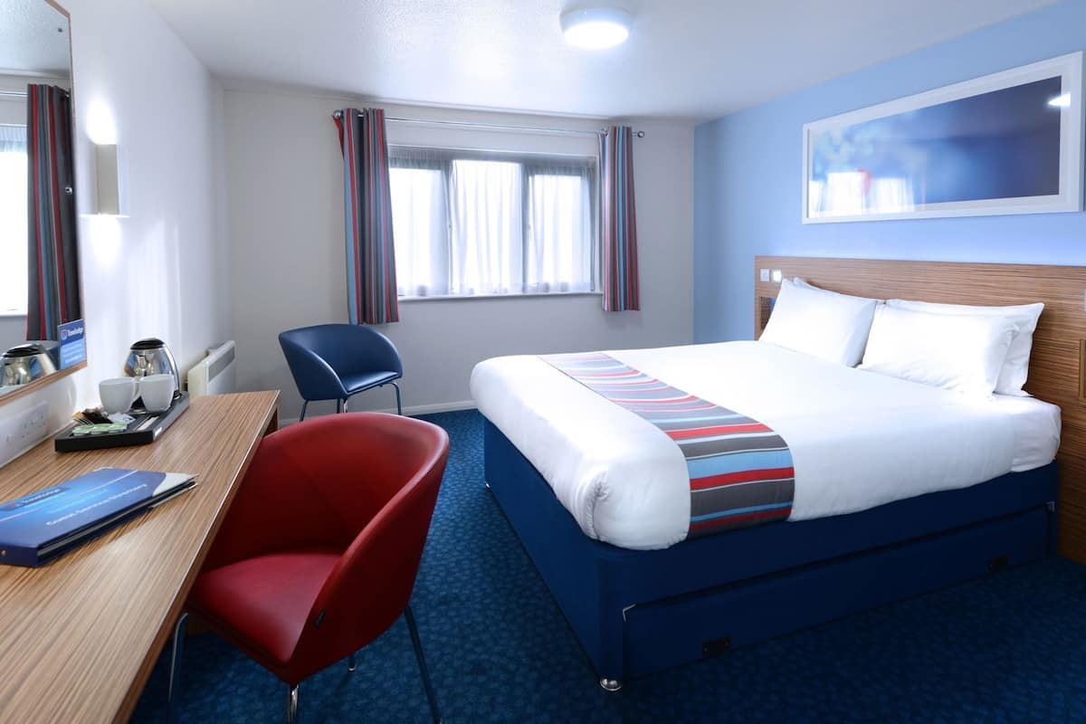 Ein Hotelzimmer mit Bett, Schreibtisch mit Stuhl, Fenster mit Vorhängen und einem an der Wand befestigten Fernseher.