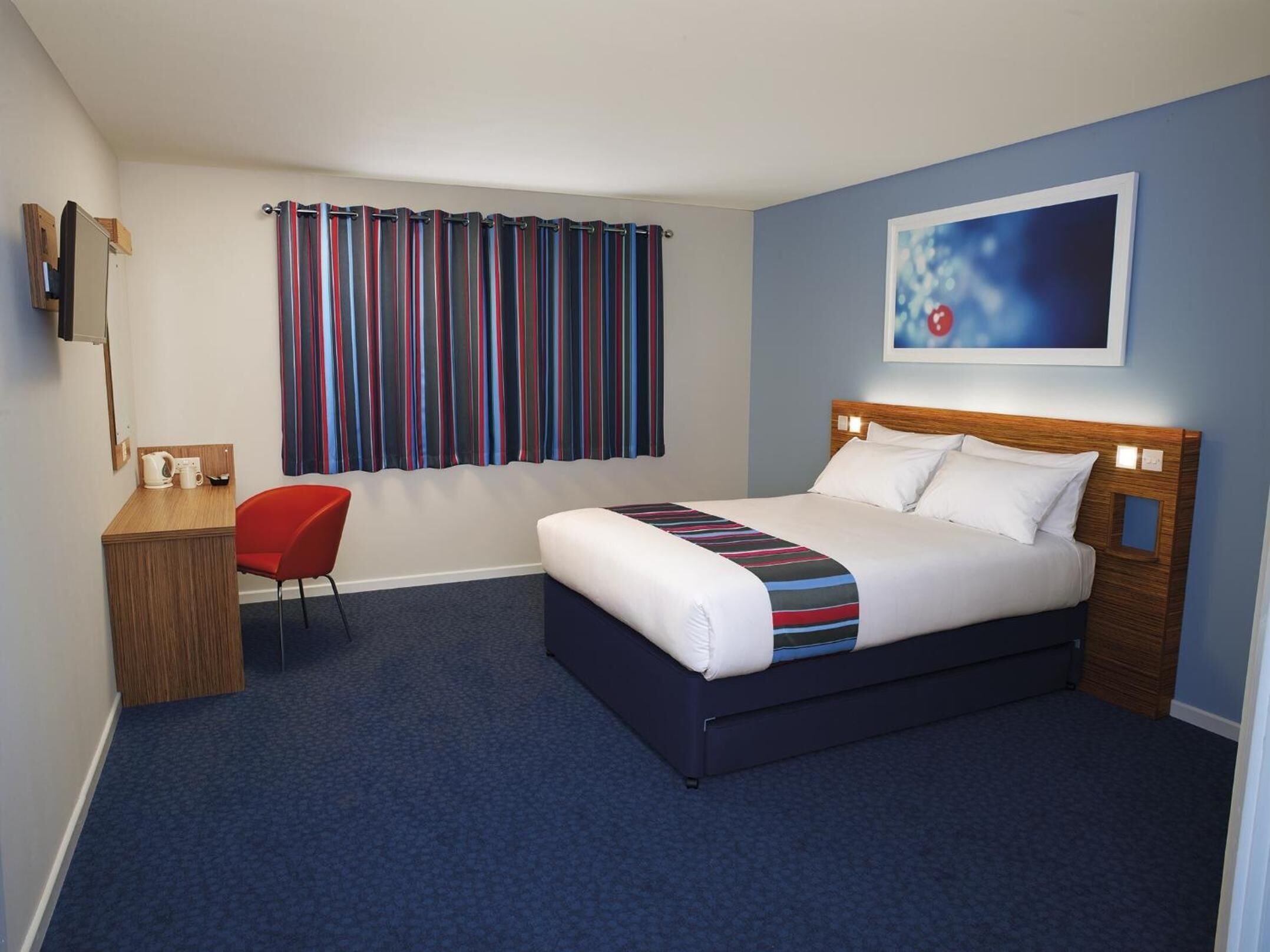 A hotel room with a bed, a desk, a chair, a window with striped curtains, and a painting on the wall.