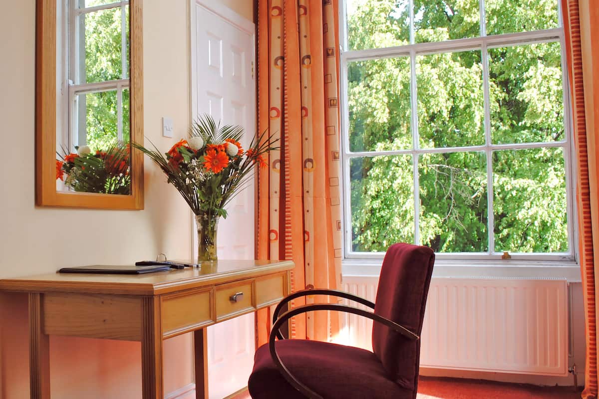 A room with a wooden desk, a chair, a mirror, a window with curtains, and a vase with flowers.