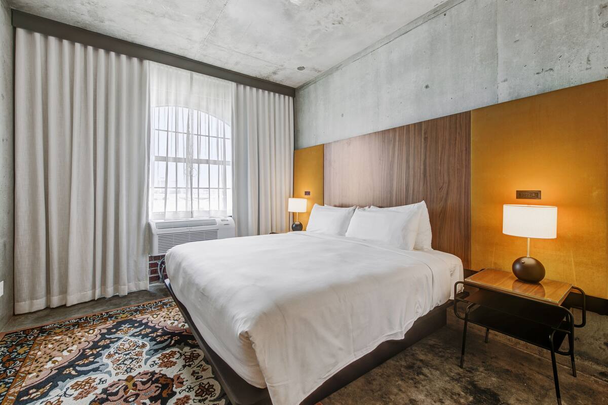 Une chambre d’hôtel avec un grand lit, une tête de lit en bois, une table de chevet avec une lampe, une fenêtre avec des rideaux et un tapis à motifs.