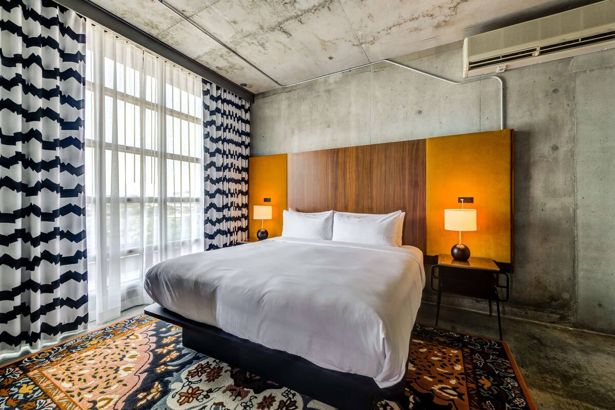 Une chambre d’hôtel moderne avec un grand lit, deux lampes de chevet, un tapis à motifs et une grande fenêtre avec des rideaux légers.