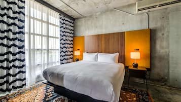 A modern hotel room with a large bed, two bedside lamps, a patterned rug, and a large window with sheer curtains.