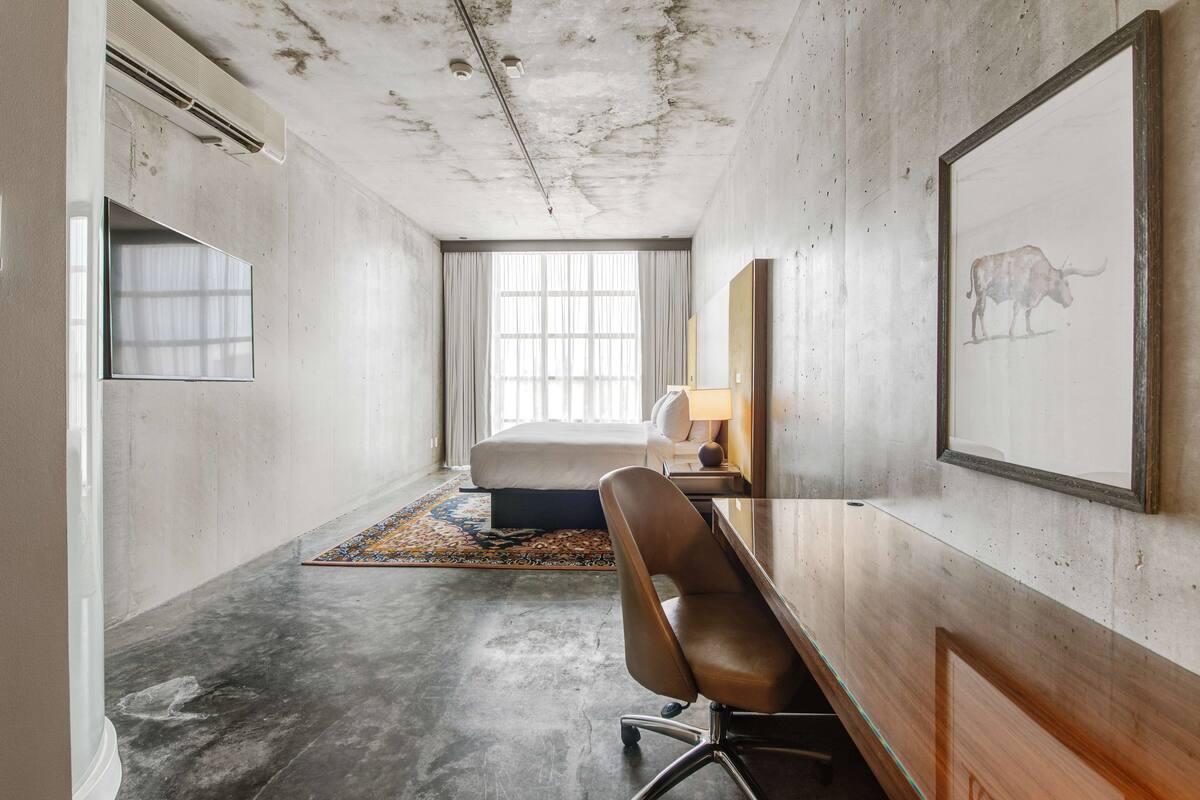 Une chambre d’hôtel comprenant un lit, un bureau, une chaise, une fenêtre avec des rideaux, un tableau encadré et un tapis à motifs.
