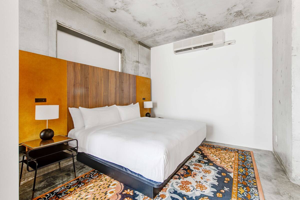 Une chambre d’hôtel avec un lit, des tables de chevet, des lampes, un tapis à motifs et un plafond en béton.