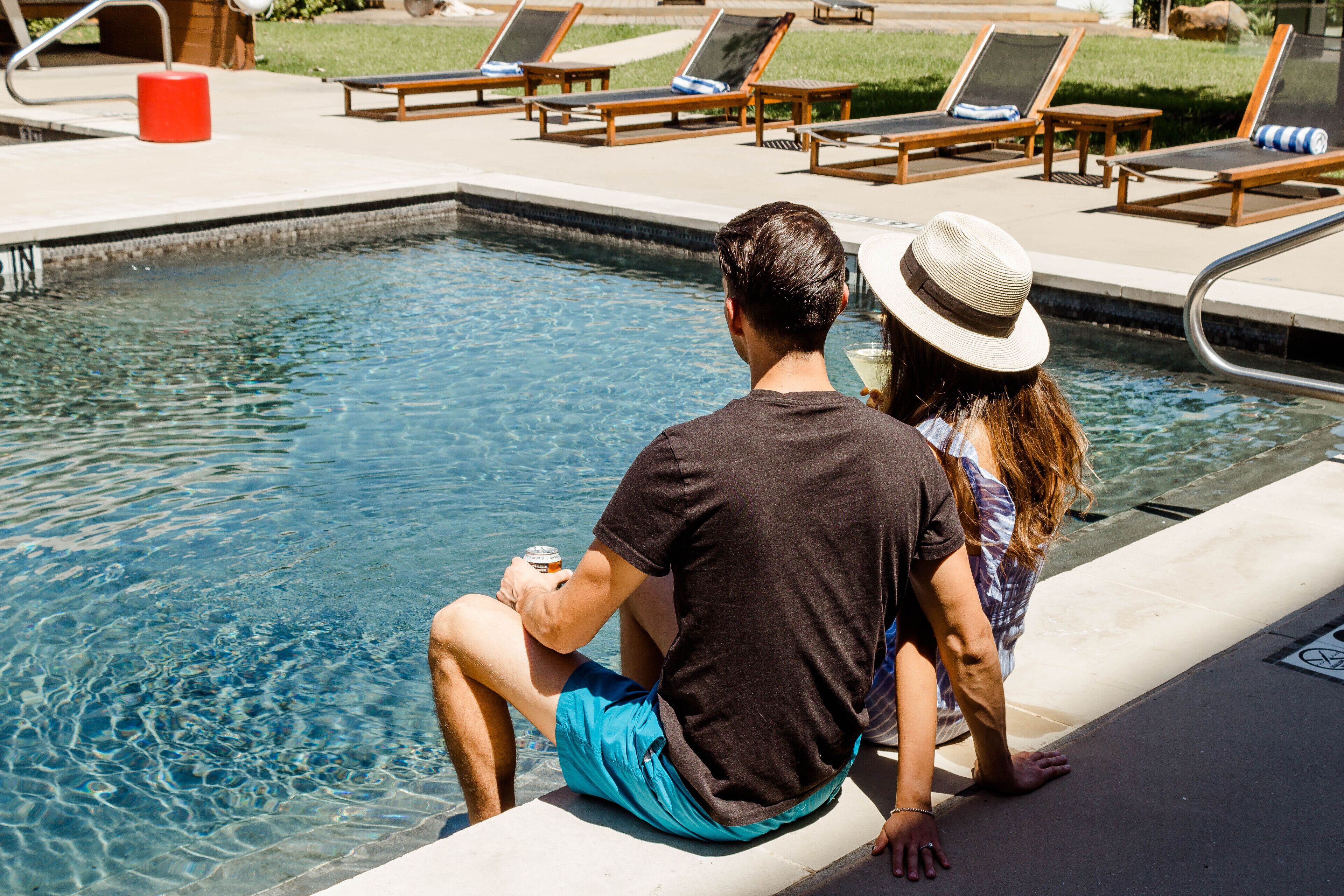 Un hombre y una mujer sentados junto a una piscina, con sillones de descanso y un cubo rojo al fondo.