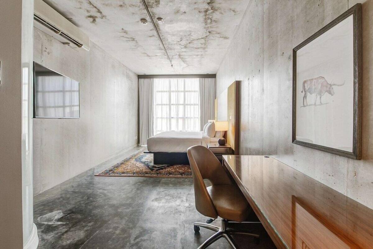 A hotel room with a bed, a desk, a chair, a window with curtains, and a framed picture on the wall.