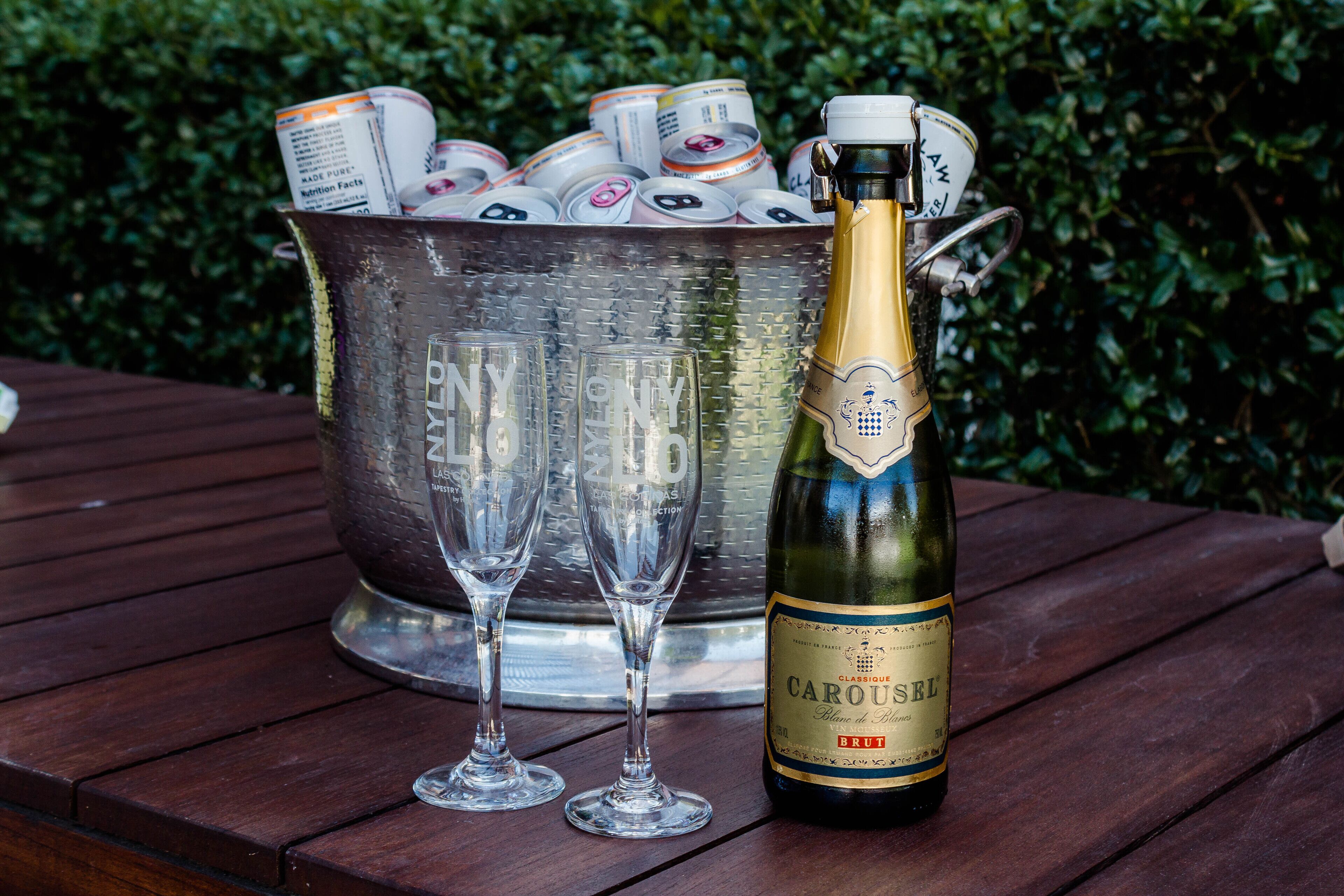 Una botella de champán Carousse Brut, dos copas vacías y un cubo de metal lleno de latas de bebidas energéticas sobre una mesa de madera.