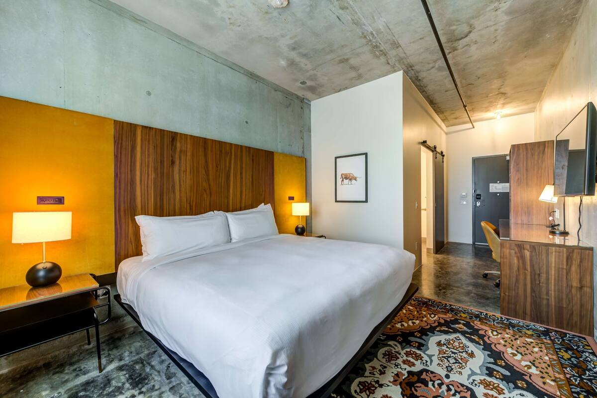 Une chambre d’hôtel avec un grand lit, deux lampes de chevet, une tête de lit en bois, un tableau encadré au mur, un bureau avec une lampe et un tapis à motifs.