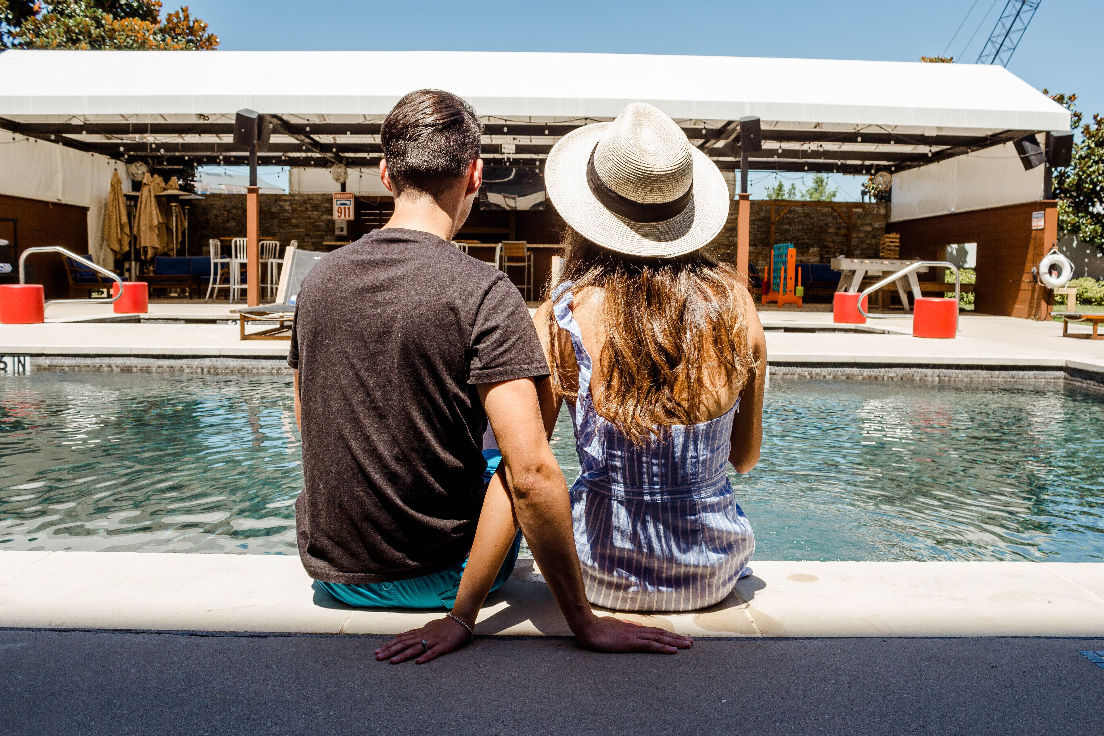 A couple sitting by a pool with a covered seating area and a sign indicating a 911 emergency phone.