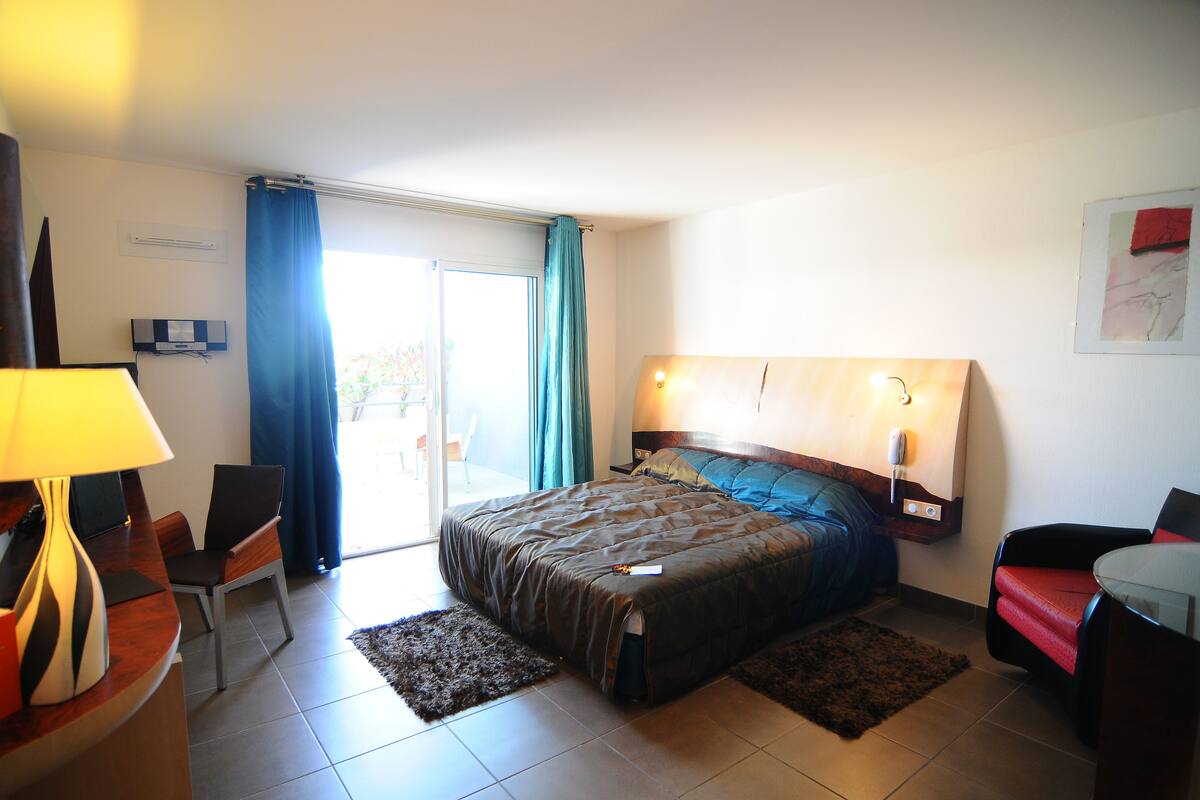 A hotel room with a bed, a desk, a chair, a lamp, and a view of the outdoors.