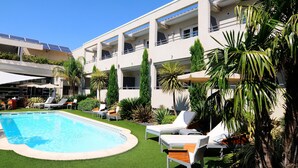 A swimming pool with sun loungers and palm trees, surrounded by a building with balconies and solar panels.