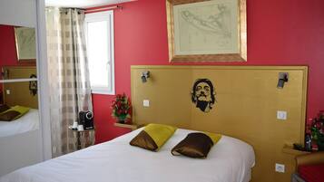 Una habitación de hotel con cama, cabecera con diseño facial, ventana con cortinas, mesita de noche con lámpara y un cuadro enmarcado en la pared.