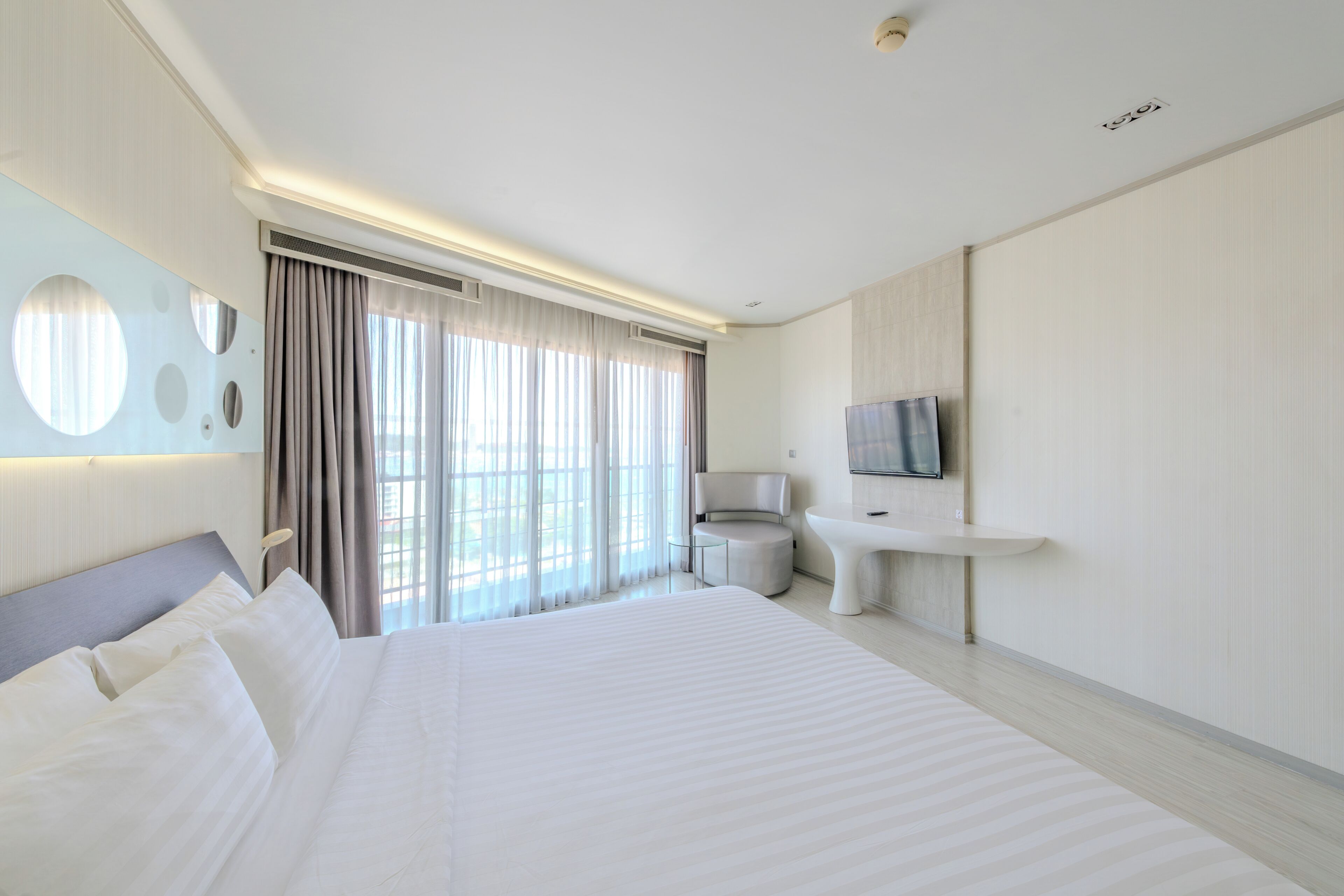 A modern hotel room with a large bed, a round mirror, a television, a round table, and a view of the outdoors.