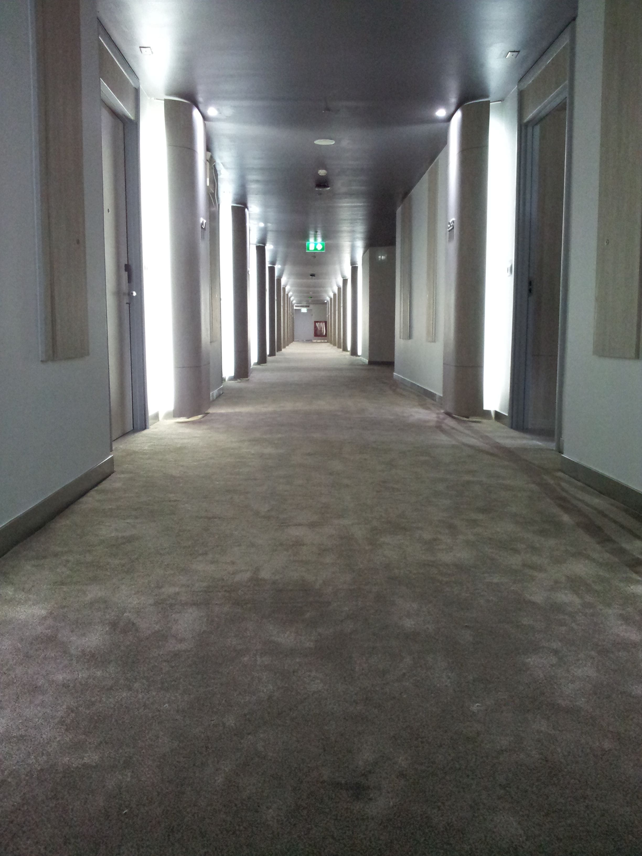 A long, narrow hallway with carpeted floors and light-colored walls, featuring doors on both sides and a green exit sign on the ceiling.