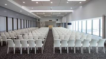 Un salón de conferencias con filas de sillas blancas, piso alfombrado y amplias ventanas con vista a la ciudad.