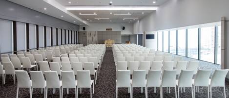Un salón de conferencias con filas de sillas blancas, piso alfombrado y amplias ventanas con vista a la ciudad.