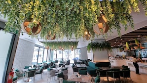 Un restaurante con plantas colgantes en el techo, mesas con cojines verdes y vistas a la ciudad a través de amplias ventanas.