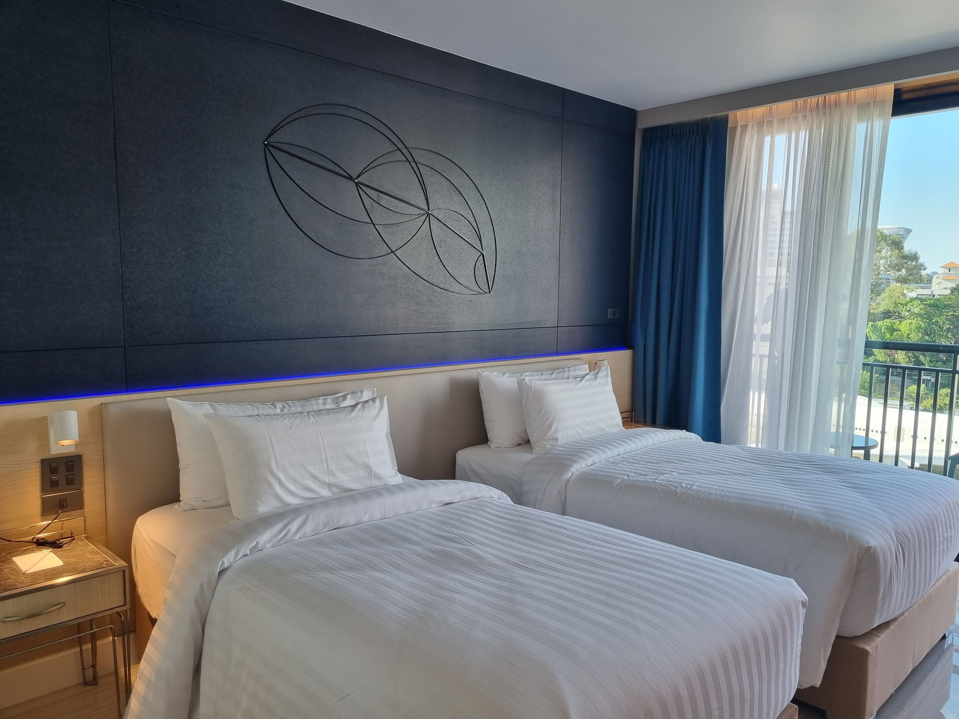 A hotel room with two beds, a modern wall art piece, a bedside table with a lamp, and a view of the outdoors through large windows.