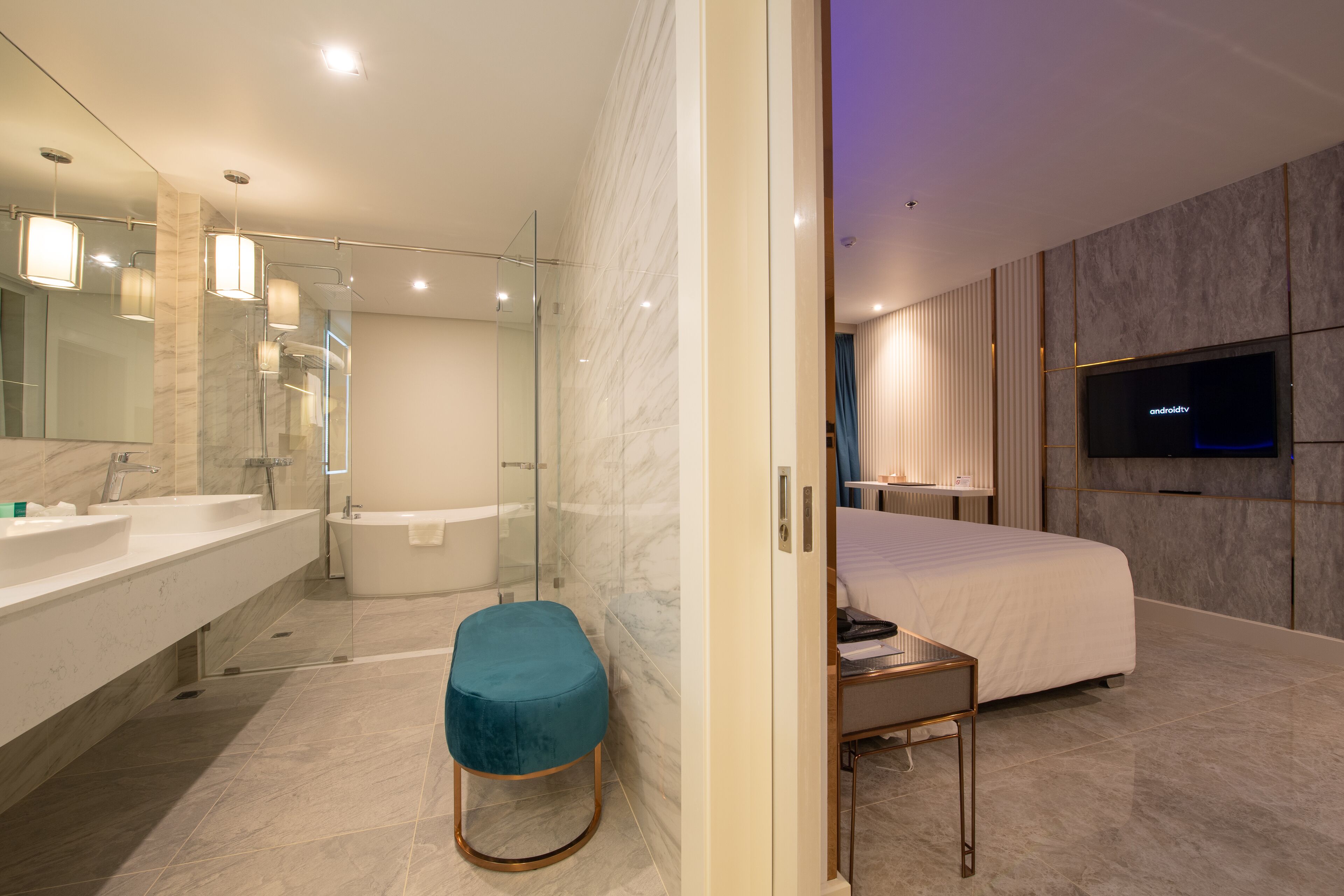 A modern hotel room with a large bed, a TV mounted on the wall, a bathroom with a glass shower enclosure, and a vanity with a mirror.