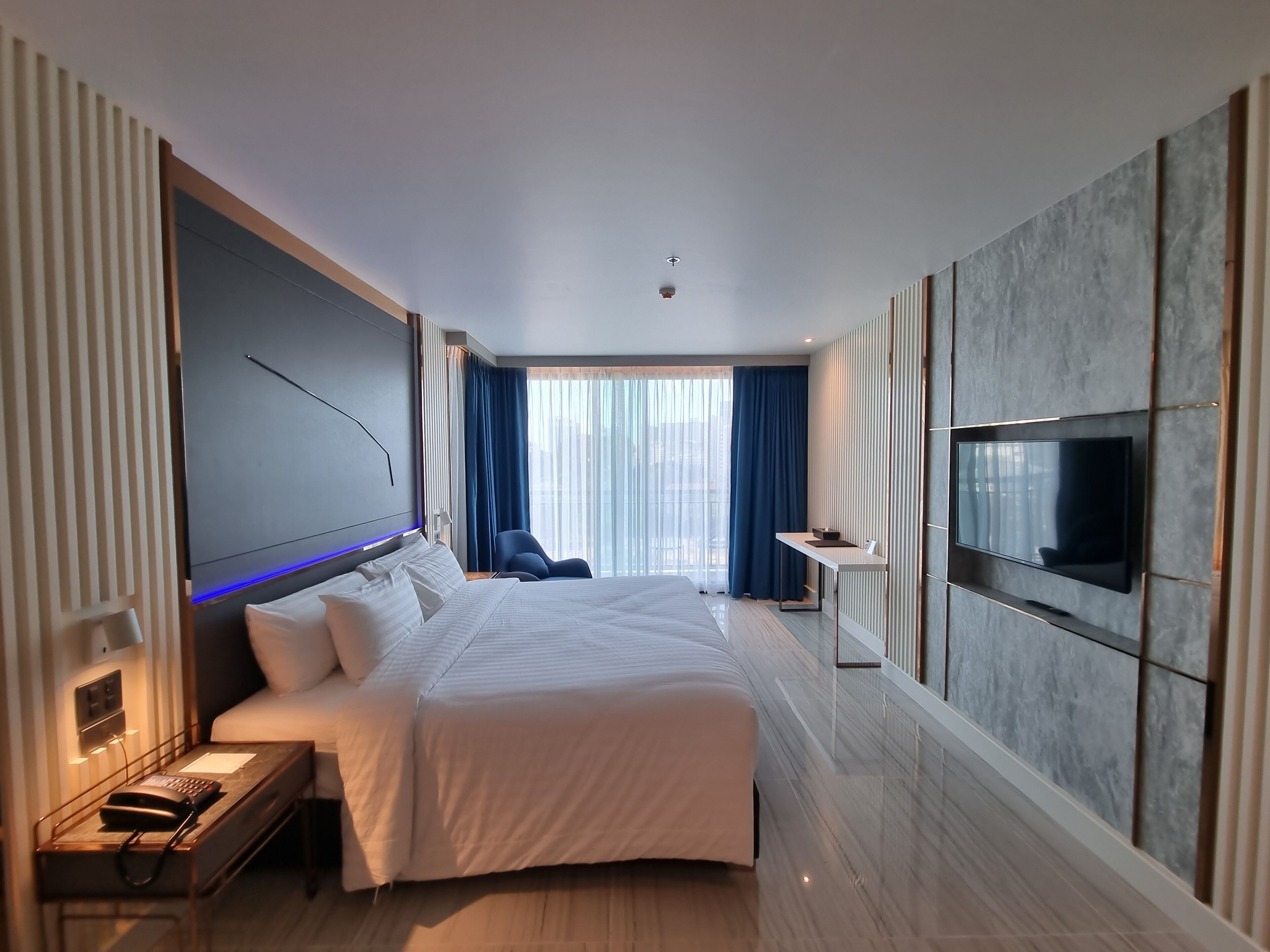 A modern hotel room with a large bed, a desk, a flat-screen TV mounted on the wall, and a view of the city through large windows.