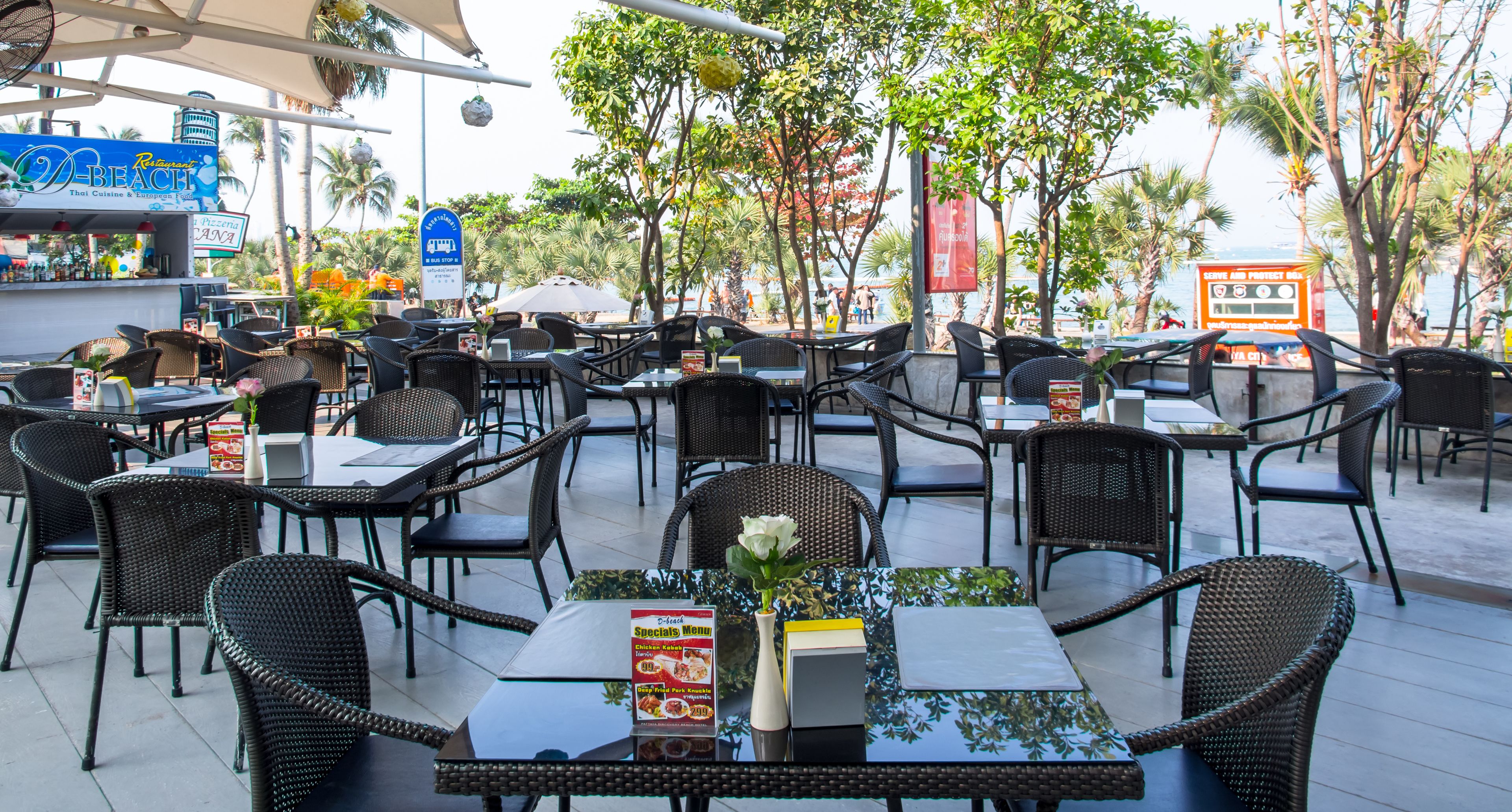 An outdoor dining area with wicker tables and chairs, a menu stand, and a view of the beach.