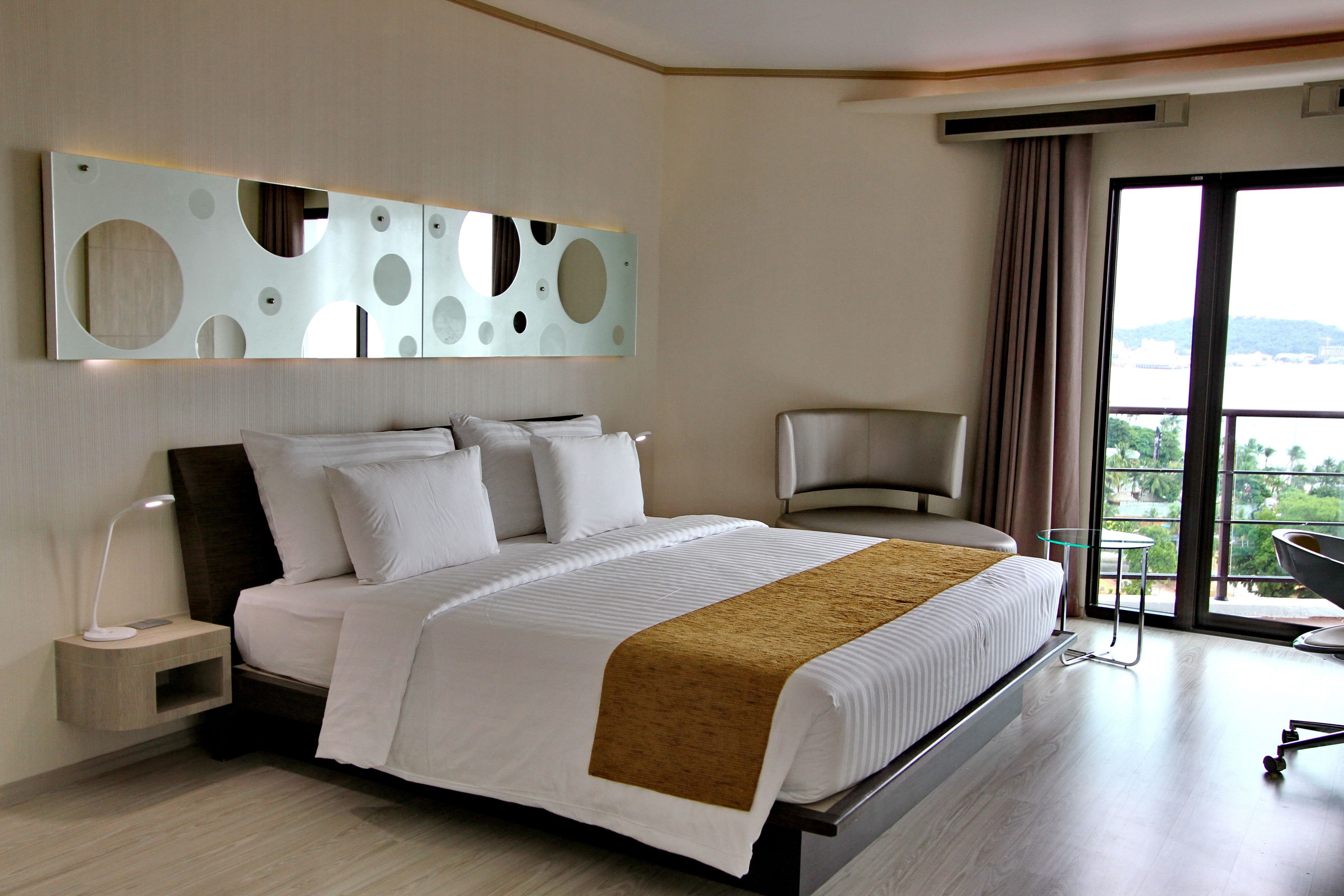 A modern hotel room with a large bed, a side table with a lamp, a chair, and a view of the outdoors through the window.